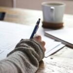 Home A hand writing with a pen beside a notebook and mug